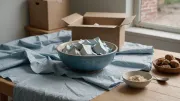 Carton d'expédition ouvert près d'une fenêtre, papier de soie froissé et bol en grès émaillé bleu posé sur la table en bois