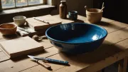 Grand bol en grès émaillé bleu sur une table d'atelier en bois, entouré de petites poteries, de feuilles de papier et d'un couteau à manche de bois